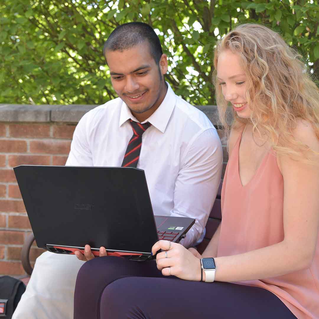 Two students, one male and one female. The mail is wearing a dress shirt and dress pants with a tie. The female student is wearing a sleeveless top and black pants.