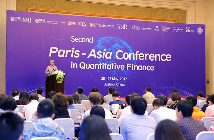 The speaker stands at a podium with flowers draped over the front edge. Behind the speaker is a large screen that says &quot;Second Paris-Asia Conference in Quantitative Finance, 26 - 27 May 2017; Suzhou, China&quot; in large white font. There are many people in the audience sitting in white chairs.